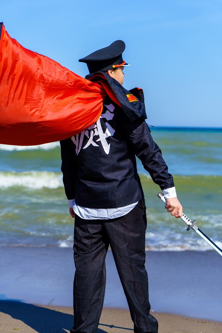 A cosplayer in a peaked cap and red-lined cape on the shore of Lake Ontario