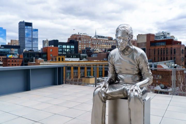 A chrome statue of a seated man on a rooftop terrace, with city buildings behind