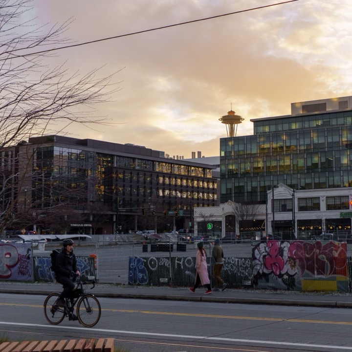 The Space Needle at sunset behind city buildings, with a cyclist passing