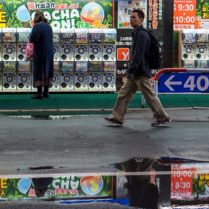 People walking past capsule toy machines, reflected in a puddle