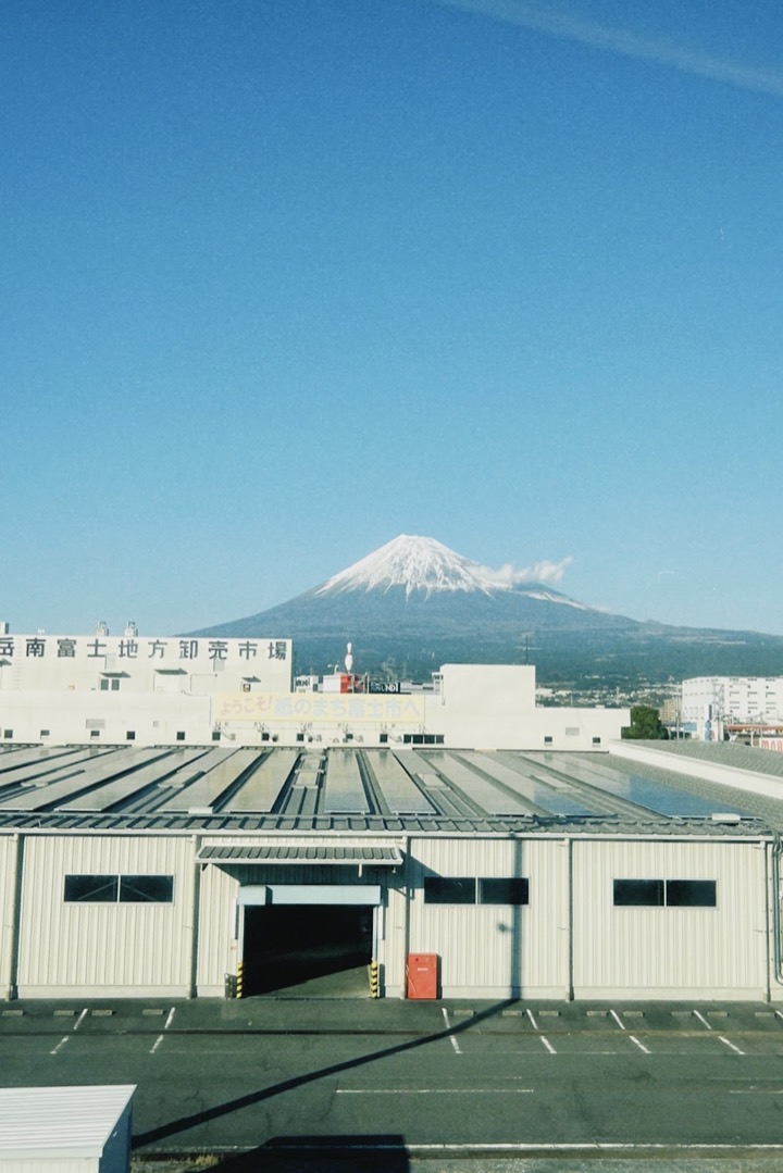 Mount Fuji rising behind a factory roof, seen from the train
